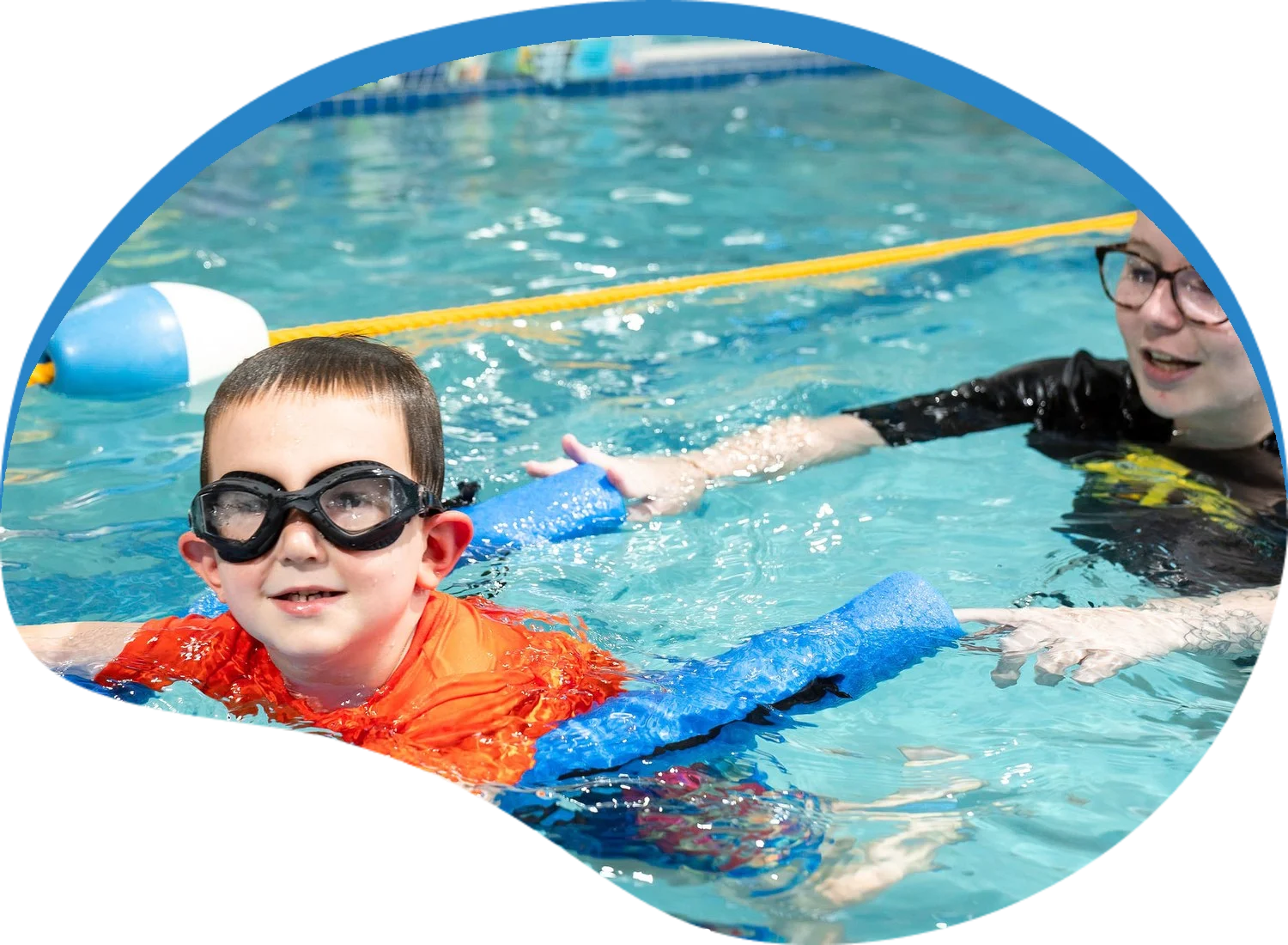 Preschooler in goggles kicking on a pool noodle with his instructor during a swim lesson at Little Fins Swim School